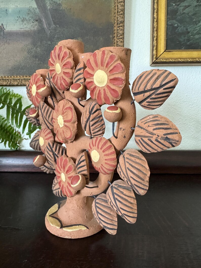 Pu&ograve; includere: Un candelabro in terracotta a forma di albero, caratterizzato da fiori dipinti a mano in rosso e giallo, foglie a righe marrone scuro e piccoli boccioli rotondi. La scultura ha una base circolare e due portacandele in cima, finitura opaca.