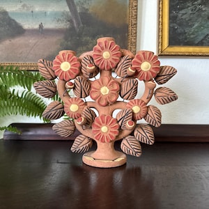 Pu&ograve; includere: Un candelabro in terracotta a forma di albero con cinque fiori rossi dai centri gialli. La scultura presenta numerose foglie marroni con dettagli incisi e piccoli accenti rotondi, il tutto sostenuto da una base circolare.