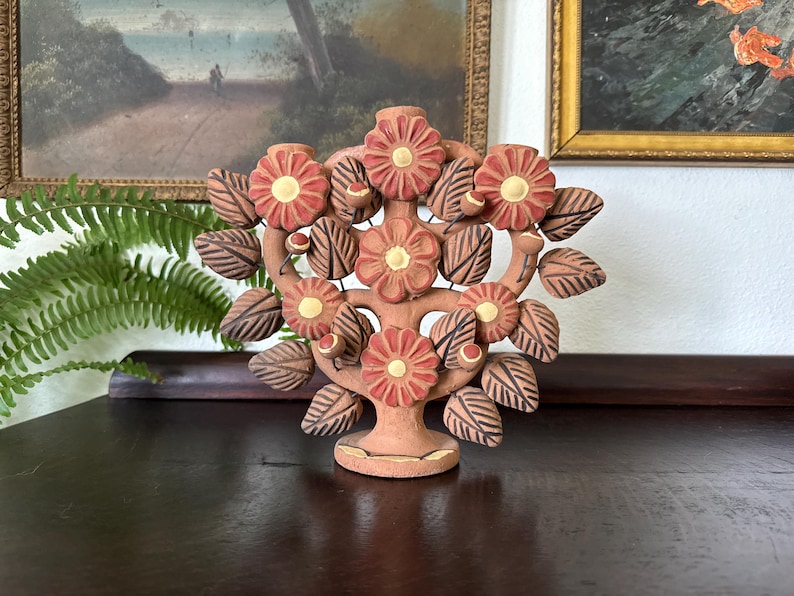 Pu&ograve; includere: Un candelabro in terracotta a forma di albero con cinque fiori rossi dai centri gialli. La scultura presenta numerose foglie marroni con dettagli incisi e piccoli accenti rotondi, il tutto sostenuto da una base circolare.