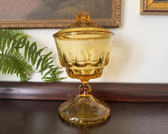 May include: A vintage amber glass candy dish with a matching lid, featuring a pedestal base and a scalloped rim design. The glass has a translucent golden hue and a smooth, reflective surface, standing on a circular footed base.