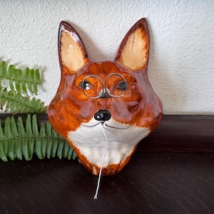 May include: A ceramic fox head wall plaque featuring a glossy orange and white finish, pointed ears, and a black nose. The fox is wearing thin, circular wire-rimmed spectacles and has a single white string hanging from its snout area.