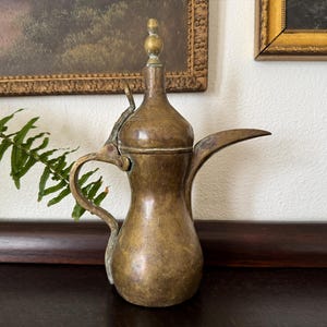 May include: A vintage brass coffee pot with a long spout and curved handle. The pot has a bulbous body and a decorative finial on the lid. The pot is displayed on a dark wooden surface.