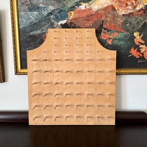 May include: A light-colored wooden display board featuring a grid of evenly spaced wooden pegs. The board has a flat rectangular base with a curved, arched top section. The surface is smooth and unfinished, designed for organizing or hanging items.