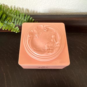 May include: A square, peach-colored plastic jewelry box featuring a circular lid with a raised relief design. The relief depicts a seated figure surrounded by swirling floral motifs and decorative scrollwork. The box has a smooth, glossy finish.