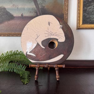 May include: A circular ceramic sculpture featuring a stylized cat design in cream and dark brown tones. The cat is depicted in a curled position with etched line details. The piece has a central circular cutout and rests on a small wooden stand.
