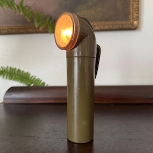 May include: An olive green vintage flashlight with a curved head and a bright, glowing light. The cylindrical body has a clip on the side. The background includes a dark wooden surface and framed artwork.