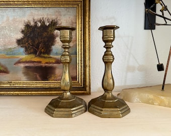 May include: A pair of matching brass candlesticks featuring a polished metallic finish, a slender central stem with decorative rings, and a wide, octagonal base. The candlesticks have a smooth, reflective surface and a classic, symmetrical shape.