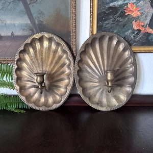 May include: A pair of oval brass wall sconces featuring a scalloped shell design with a beaded edge. Each sconce has a central candle holder cup attached to the metal plate. The brass has a warm, aged metallic finish with visible patina.