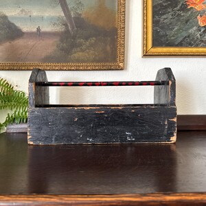 May include: A rectangular wooden tool caddy with a distressed black finish and visible wood grain. It features two high side panels and a horizontal carrying handle across the top, which is painted black with small, evenly spaced red accents.