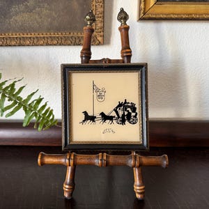 May include: A black and white silhouette image of a horse-drawn carriage with passengers, framed in a black frame with a gold trim. The frame is on a wooden easel.