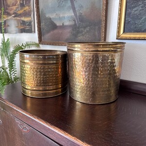 May include: Two cylindrical brass planters with a hammered texture. The smaller planter on the left features horizontal bands near the top and bottom, while the larger planter on the right has a smooth rim and a consistent hammered finish throughout.