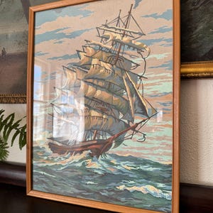 May include: A framed painting of a ship with white sails sailing on a blue ocean. The ship is in the foreground of the painting, and the ocean is in the background. The painting is in a wooden frame.