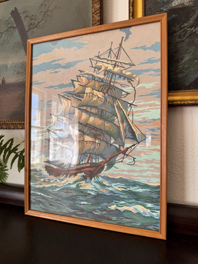 May include: A framed painting of a ship with white sails sailing on a blue ocean. The ship is in the foreground of the painting, and the ocean is in the background. The painting is in a wooden frame.
