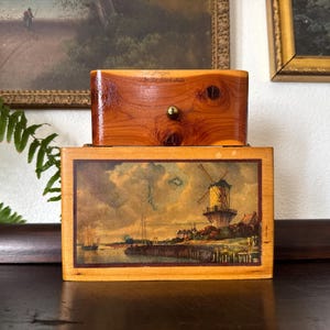 May include: A rectangular wooden box with a polished finish and a small brass knob on the top section. The front panel features a detailed landscape print of a windmill near a body of water with boats, framed by a dark border on a light wood base.