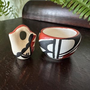 May include: Two small, hand-painted ceramic pots. One is a small, cream-colored vessel with a red rim and black and red geometric designs. The other is a small, cream-colored pot with a red rim and black and red geometric designs.