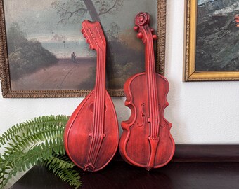 May include: Two wall-mounted decorative musical instrument plaques in a distressed red finish. One plaque is shaped like a mandolin with a long neck and rounded body, while the other is shaped like a violin with a scroll and f-holes.