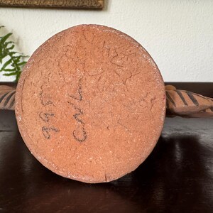 Pu&ograve; includere: Una base circolare in argilla color terracotta con una superficie ruvida e strutturata. La base presenta segni scritti a mano con inchiostro nero che riportano 995 e Carl. Due estensioni simmetriche a forma di ala con strisce marrone scuro.
