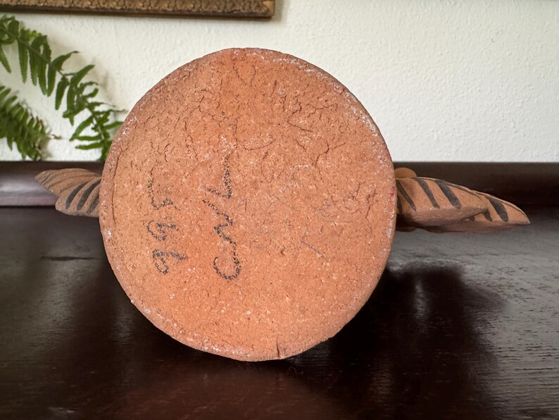 Pu&ograve; includere: Una base circolare in argilla color terracotta con una superficie ruvida e strutturata. La base presenta segni scritti a mano con inchiostro nero che riportano 995 e Carl. Due estensioni simmetriche a forma di ala con strisce marrone scuro.