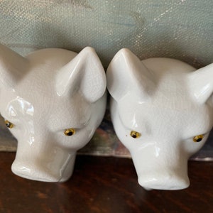 Puede incluir: Dos cabezas de cerdo de cer&aacute;mica blanca con un acabado de esmalte craquelado brillante. Cada cabeza de cerdo tiene orejas puntiagudas, un hocico y ojos peque&ntilde;os, redondos y oscuros con iris de color dorado. Las piezas est&aacute;n esculpidas con detalle.