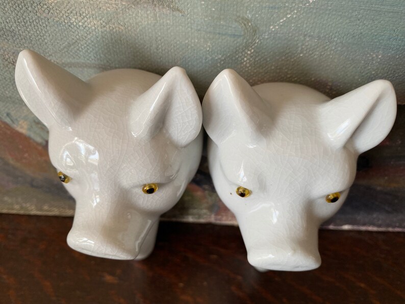 Puede incluir: Dos cabezas de cerdo de cer&aacute;mica blanca con un acabado de esmalte craquelado brillante. Cada cabeza de cerdo tiene orejas puntiagudas, un hocico y ojos peque&ntilde;os, redondos y oscuros con iris de color dorado. Las piezas est&aacute;n esculpidas con detalle.