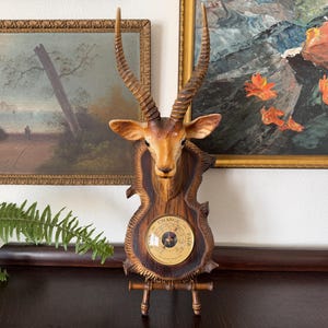 May include: A wall-mounted barometer featuring a sculpted gazelle head with long, spiraled horns. The animal head is mounted on a dark wood-grain plaque that holds a circular analog barometer dial with the text "CHANGE" and "FAIR" printed on its face.
