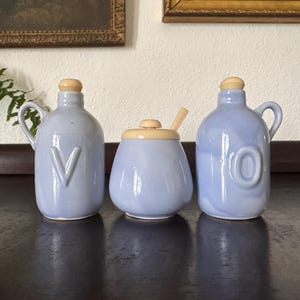 May include: A set of three light blue ceramic kitchen containers. Two bottles with wooden stoppers and handles feature the letters "V" and "O". A round jar with a wooden spoon and lid completes the set. The containers are on a dark surface.