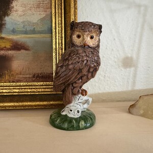 May include: A brown ceramic owl figurine with detailed feather textures, perched atop a small white and green base. The owl has large, expressive eyes and is set against a backdrop of a framed landscape painting.