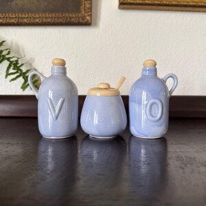 May include: Three light blue ceramic containers with wooden lids. Two have handles and embossed letters "V" and "O". The center container is a sugar bowl with a wooden spoon. The containers are on a dark wooden surface.