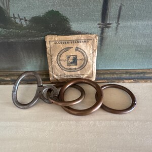 May include: A vintage metal bull ring set featuring a dark, weathered finish. The set includes a hinged, oval-shaped tool and two circular rings. Behind the metal pieces sits a square, tan paper envelope labeled Allover-Standard Bull Ring.