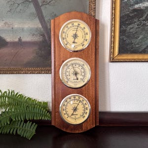May include: A vertical wooden weather station featuring three circular gauges with cream-colored faces and silver rims. The top gauge measures humidity, the middle gauge is a barometer labeled Rain, Change, and Fair, and the bottom gauge measures temperature.