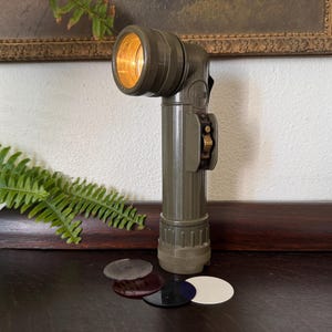 May include: An olive-drab vintage military flashlight with a pivoting head and a switch. The light is illuminated, showing a warm glow. Several colored lenses are at the base. A fern and framed artwork are in the background.