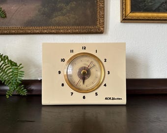 May include: A cream-colored rectangular RCA Victor clock with a central circular dial featuring black numerals from one to twelve. The clock face is encased in a gold-toned circular frame with thin black hands and a small red second hand.