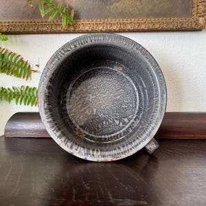 May include: A round, deep metal basin featuring a gray and white mottled enamel finish. The interior displays a distinct radial pattern, while the exterior rim shows signs of wear with small patches of rust visible along the edges of the metal.