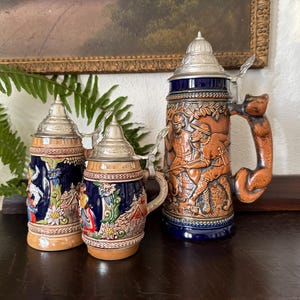 May include: Three decorative beer steins with detailed relief designs. The steins feature silver lids and handles, with the largest stein showcasing a brown handle shaped like a fox. The steins are in shades of blue, brown, and tan, with intricate scenes.