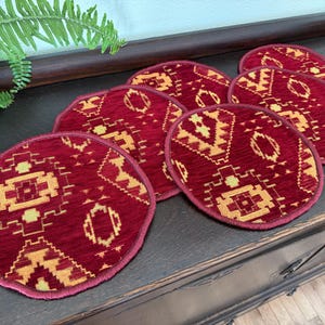 May include: Set of seven round coasters with a burgundy red background and a geometric pattern in gold and beige. The coasters are arranged on a dark wooden surface, showcasing their design and color.