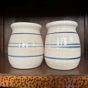 May include: Two white ceramic jars with blue stripes. The jars are round and have a slightly rounded bottom. They are sitting on a wooden shelf.