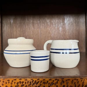 May include: Three ceramic kitchenware items with blue stripes. A white pitcher, a white jar with a lid, and a white cup.