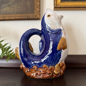 May include: A blue and white ceramic pitcher shaped like a fish. The fish has a brown tail fin and is decorated with a blue and white pattern. The pitcher is sitting on a dark brown surface.