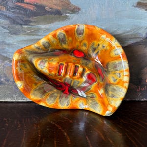 May include: A vintage ceramic ashtray with a unique, organic shape. The ashtray features a vibrant color palette of orange, yellow, and red, with a glossy finish. It has a central area for holding cigarettes and a curved design.