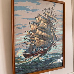May include: A framed painting of a large sailing ship with white sails against a blue sky and ocean. The ship is in the foreground and the sky and ocean are in the background. The painting is in a wooden frame.