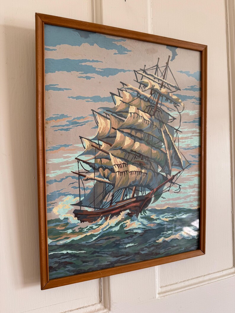 May include: A framed painting of a large sailing ship with white sails against a blue sky and ocean. The ship is in the foreground and the sky and ocean are in the background. The painting is in a wooden frame.