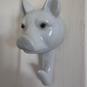 Puede incluir: Un gancho de pared de cer&aacute;mica blanca con forma de cabeza de cerdo. El cerdo presenta un acabado de esmalte brillante y agrietado, orejas puntiagudas y dos ojos peque&ntilde;os, redondos y de color &aacute;mbar. El gancho se extiende desde la barbilla.