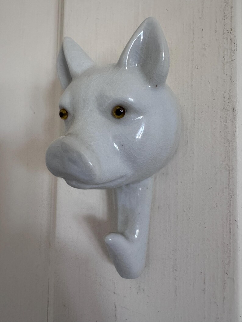 Puede incluir: Un gancho de pared de cer&aacute;mica blanca con forma de cabeza de cerdo. El cerdo presenta un acabado de esmalte brillante y agrietado, orejas puntiagudas y dos ojos peque&ntilde;os, redondos y de color &aacute;mbar. El gancho se extiende desde la barbilla.