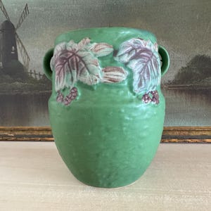 May include: A green ceramic vase with a textured surface and a design of leaves and berries in shades of purple and green. The vase has two handles.