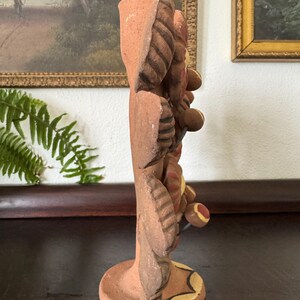Pu&ograve; includere: Un portacandele in terracotta caratterizzato da uno stelo verticale decorato con motivi scultorei a forma di foglie e fiori circolari. La superficie in argilla presenta toni naturali della terra con accenti dipinti in giallo e rosso.