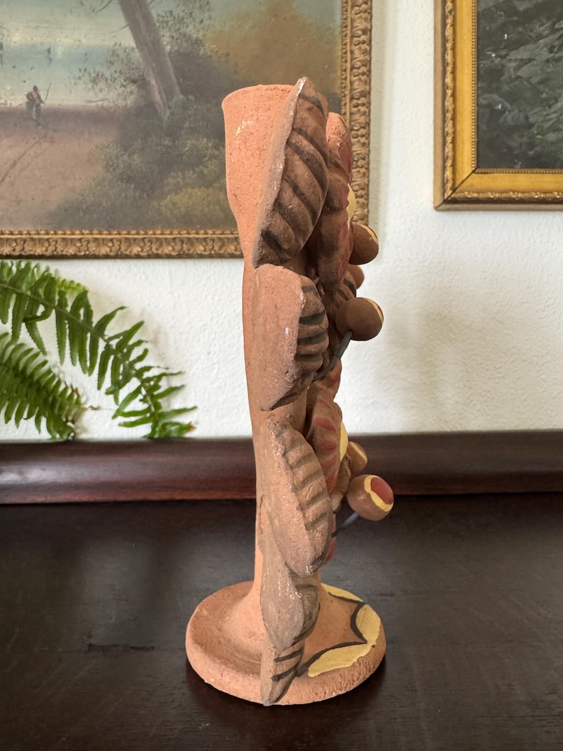 Pu&ograve; includere: Un portacandele in terracotta caratterizzato da uno stelo verticale decorato con motivi scultorei a forma di foglie e fiori circolari. La superficie in argilla presenta toni naturali della terra con accenti dipinti in giallo e rosso.