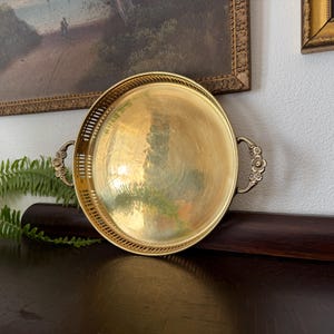 May include: A round, gold-toned metal tray with decorative handles. The tray has a polished surface and a detailed, pierced border. The handles feature floral designs. The tray is displayed on a dark wooden surface.