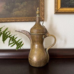 May include: A vintage brass coffee pot with a long spout, curved handle, and a decorative lid. The pot has a tarnished, aged appearance, suggesting it is an antique. The background includes framed artwork and a fern.