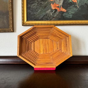 May include: A handcrafted wooden tray with an octagonal shape. The tray is constructed from numerous small wooden pieces, creating a textured surface. A red base supports the tray, which is displayed on a dark wooden surface.