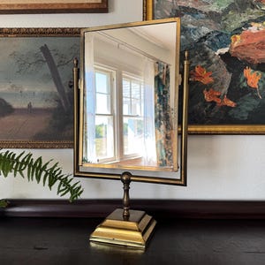 May include: A vintage brass vanity mirror with a rectangular frame and a square base. The mirror is set against a backdrop of framed artwork and a dark wooden surface. The mirror reflects a window with white trim and curtains.